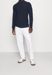 Mężczyzna w granatowym, prążkowanym swetrze z golfem, białych spodniach o podwiniętych nogawkach i białych sneakersach z czerwonymi paskami, stojący na jednolitym tle.