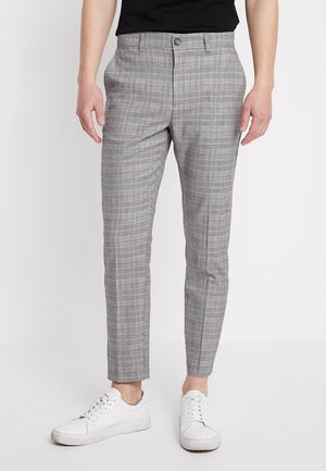 Mężczyzna ubrany w dopasowane, szare spodnie w kratę oraz białe trampki, stojący na tle jednolitego, białego tła.