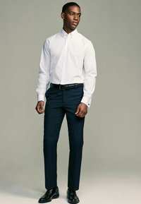 Weißes Hemd mit langen Ärmeln, dunkelblaue Hose, schwarzer Gürtel und glänzende schwarze Schuhe; glattes Gewebe und taillierte Passform.