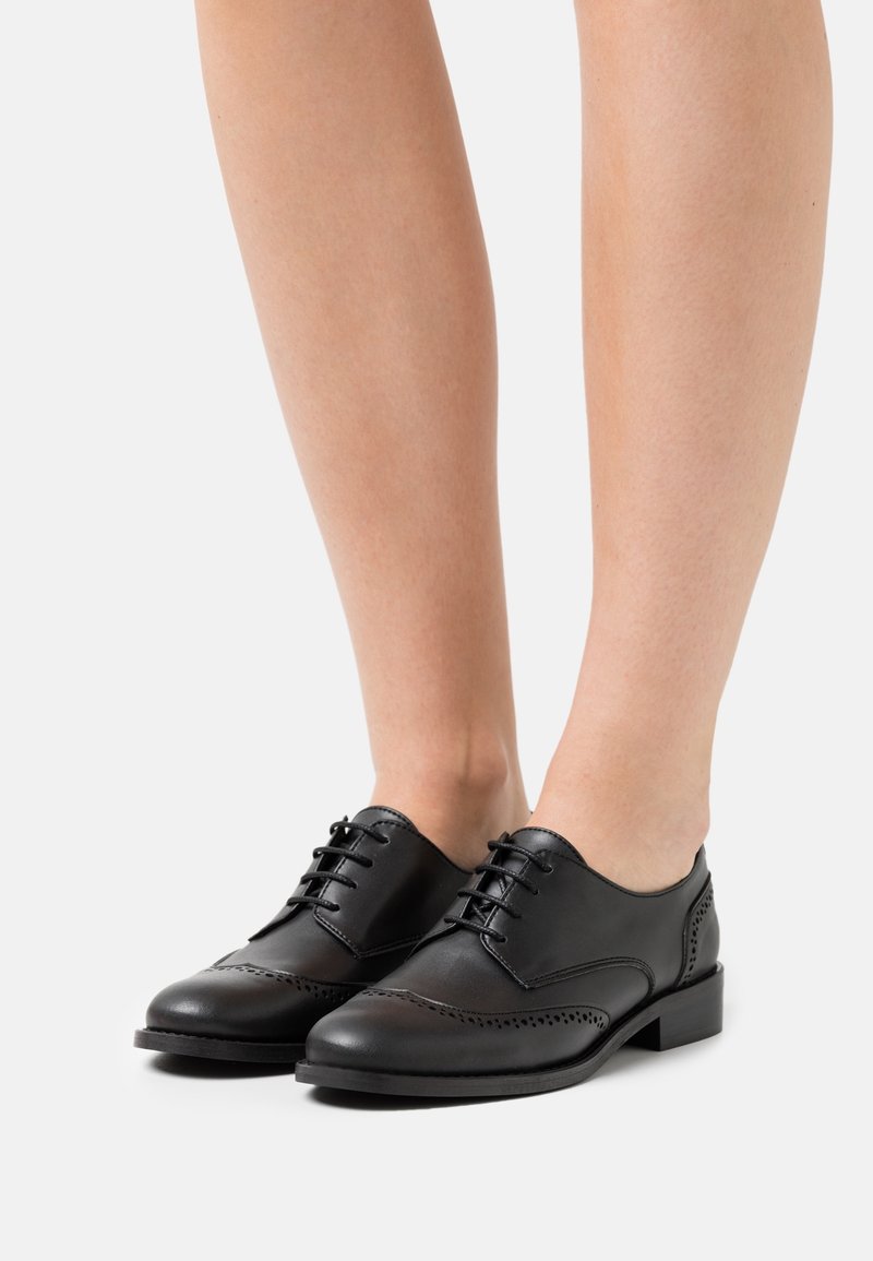 Chaussures brogues noires en cuir, à lacets, portées pieds nus, avec des perforations décoratives et des talons bas, sur un fond clair uni.
