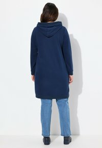 Felpa lunga con cappuccio di colore blu navy, maniche lunghe, polsini e orlo a costine. Indossata con jeans azzurri e scarpe con plateau nere.