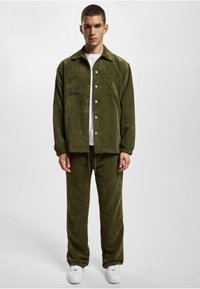 Olive green corduroy jacket with button front and two pockets, paired with matching pants. White t-shirt and sneakers complete the look.