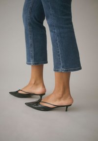 Jambes vêtues d’un jean bleu coupé court et de mules noires à talons aiguilles bas avec fines brides sur un fond neutre.