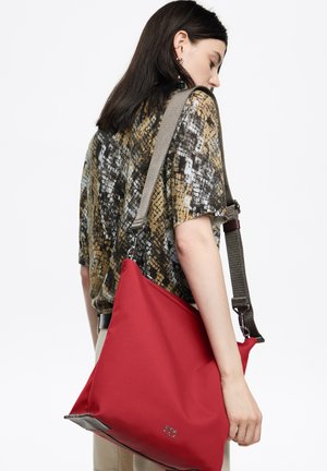 Kobieta w wzorzystej koszuli niesie dużą czerwoną torebkę na ramię z szerokim szarym paskiem na białym tle.