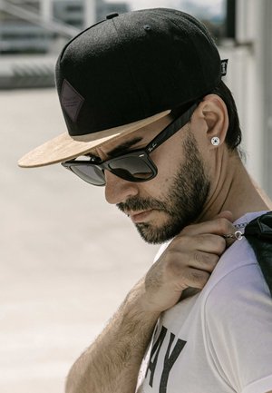 Black snapback hat with a light tan brim, dark sunglasses, white t-shirt with text, and a leather bag strap held by a hand with short hair.