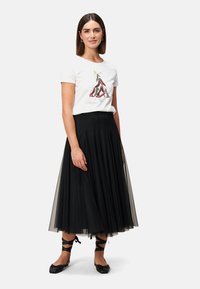 T-shirt blanc avec un graphisme de chaussures, assorti à une jupe en tulle noire. La jupe présente des plis et arrive au niveau des mollets. Ballerines noires avec des lanières à la cheville.