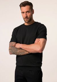 Zwarte katoenen T-shirt met korte mouwen, een ronde halslijn en een aansluitende zoom. Model heeft zichtbare tatoeages op de armen. Eenvoudig, effen ontwerp.