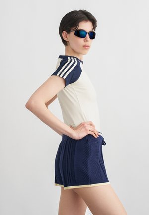 Persoon met donkere zonnebril, een crème- en marineblauw kortermig shirt met strepen, en marineblauwe gebreide korte broek, die poseert met handen in de zij.