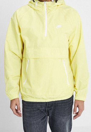 Personne portant un coupe-vent Nike jaune clair avec une poche zippée à l'avant et des détails de zip blancs sur les manches et la capuche.