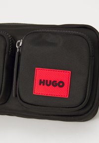 Schwarze Nylon-Tasche mit mehreren Reißverschlussfächern und einem auffälligen roten und schwarzen "HUGO" Logo-Patch. Die strukturierte Oberfläche verbessert den Halt.