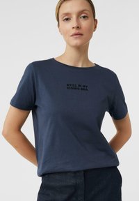 Granatowy t-shirt z krótkim rękawem wykonany z miękkiego materiału, z okrągłym dekoltem, z czarnym napisem "WCIĄŻ W MOJEJ IKONICZNEJ EPOCE" wyśrodkowanym na przodzie.