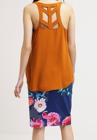 Top arancione senza maniche con schiena incrociata, abbinato a una gonna a lunghezza ginocchio con fantasia floreale che presenta fiori rosa e bianchi su uno sfondo blu navy.
