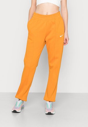 Person iført lyse orange Nike træningsbukser og farverige sneakers, stående mod en ensfarvet lys baggrund.