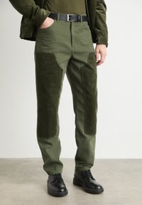 Green corduroy pants with contrasting textured patches, featuring a straight fit and black belt, paired with black shoes on a neutral background.