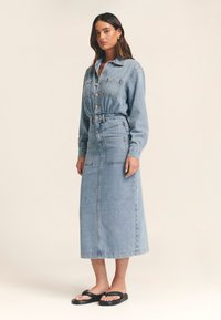 Vaaleansininen denimpuku, jossa on etunapit, kaulus, pitkät hihat, kaksi rintataskua ja vyötäröllä vyö. Yhdistetty mustiin sandaaleihin.