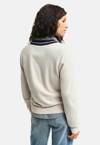 Beiger Pullover mit strukturierter Oberfläche, gerippten Bündchen und einem navygestreiften Kragen, von hinten gesehen, über einem gestreiften Hemd getragen.