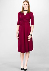 Burgundowa sukienka do kolan z dekoltem w kształcie litery V, trzyczwartowymi rękawami i zebranym talią. Gładki materiał i zwiewna spódnica; noszona z czarnymi szpilkami.
