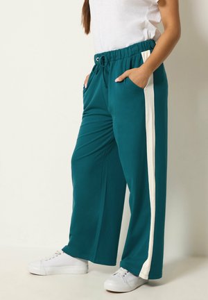Vrouw draagt turquoise wijd uitlopende broek met witte zijstrepen, wit T-shirt en witte sneakers, staat met handen in de zakken.