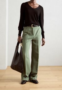 Maglione nero a scollo a V, pantaloni verde a gamba larga, borsa nera e mocassini. Presenta dettagli con bottoni sulle maniche e finiture in oro.