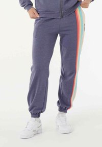 Persoon in paarse joggers met regenboogstrepen aan de zijkanten en witte sneakers, handen rustend bij de zakken.