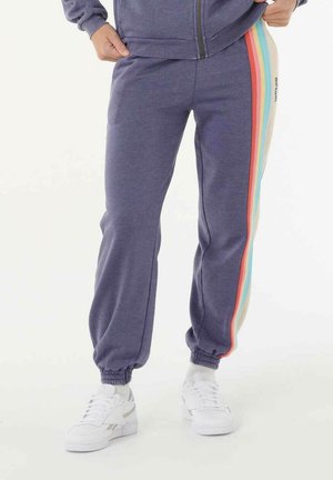 Persona que lleva pantalones deportivos morados con rayas arcoíris a los lados y zapatillas blancas, con las manos descansando cerca de los bolsillos.