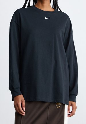 Persoon die een loszittend zwart shirt met lange mouwen draagt, met een klein wit Nike-logo op de borst en een donkere broek met gele Nike-details.