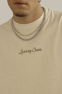 Camiseta de algodón color crema con la frase "Luxury Chaos" bordada en cursiva marrón, combinada con collares de cadena plateada en capas.