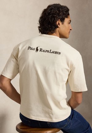 Homme aux cheveux bouclés assis sur un tabouret, portant un t-shirt crème Polo Ralph Lauren avec logo et texte au dos, associé à un jean bleu foncé.