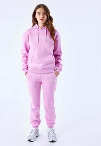 Roze hoodie en bijpassende sweatpants van een zachte stof, met een voorzak en geborduurd logo. Sneakers maken de outfit compleet.