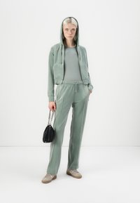 Groene velours trainingsoutfit met een ritssluiting hoodie en aansluitend top, gecombineerd met taps toelopende broek. Bevat een zwarte handtas met kettinghandvat en beige sneakers.