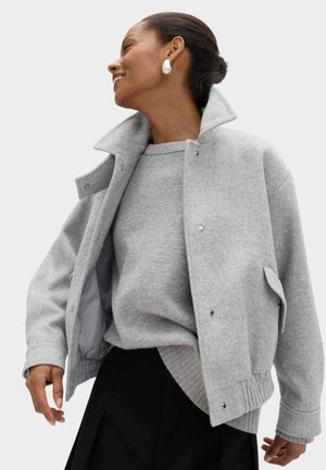 Chaqueta gris de mezcla de lana con cuello alto, cierres a presión y puños acanalados, superpuesta sobre un suéter gris a juego. Falda negra visible.