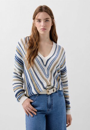 Sweter w paski z dekoltem w serek w odcieniach beżu, niebieskiego i białego, o luźnym kroju i ściągaczach na rękawach. Noszony z jasnoniebieskimi dżinsami i białym paskiem.