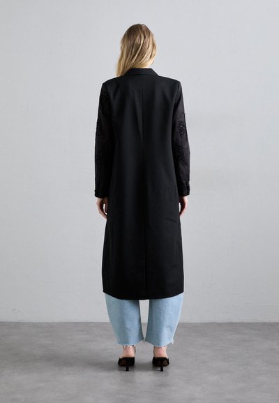 Garš melns mētelis ar izšūtām caurspīdīgām piedurknēm, taisna piegriezuma, sānu kabatas, apvienots ar gaiši zilām plašām džinsām un melniem papēžiem.