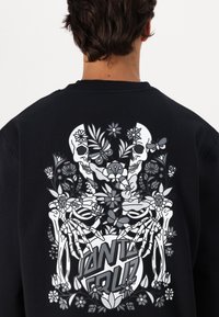 Zwarte sweatshirt met een gedetailleerde grijs-witte afbeelding van twee schedels omringd door bloemen en vlinders, met opvallende tekst in het midden.
