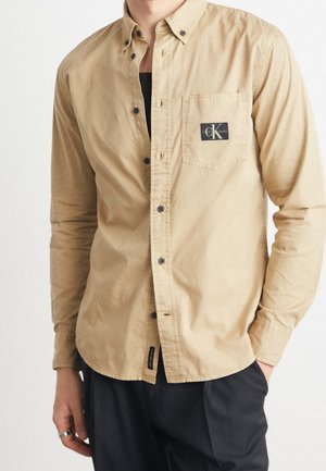 Homme portant une chemise beige boutonnée avec un logo Calvin Klein noir sur la poche poitrine et un pantalon noir, main dans la poche.