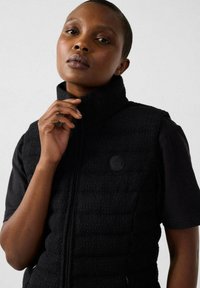 Gilet nero con texture, colletto alto e zip frontale; presenta un logo tondo sul lato sinistro. Indossato sopra una maglietta nera a maniche corte.