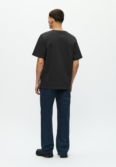 Tricou negru din bumbac, cu guler rotund, mâneci scurte și croială lejeră, purtat cu pantaloni închisi la culoare și încălțăminte neagră.