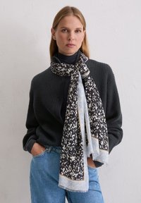 Zwart-wit sjaal met patroon en lichtblauwe accenten, gemaakt van een zacht materiaal, elegant om de nek gedrapeerd. Draagt een donker trui.