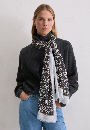 Zwart-wit sjaal met patroon en lichtblauwe accenten, gemaakt van een zacht materiaal, elegant om de nek gedrapeerd. Draagt een donker trui.