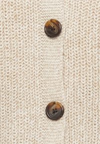 Nærbilde av beige strikket stoff med to store brune marmorerte knapper sydd i en vertikal rad.