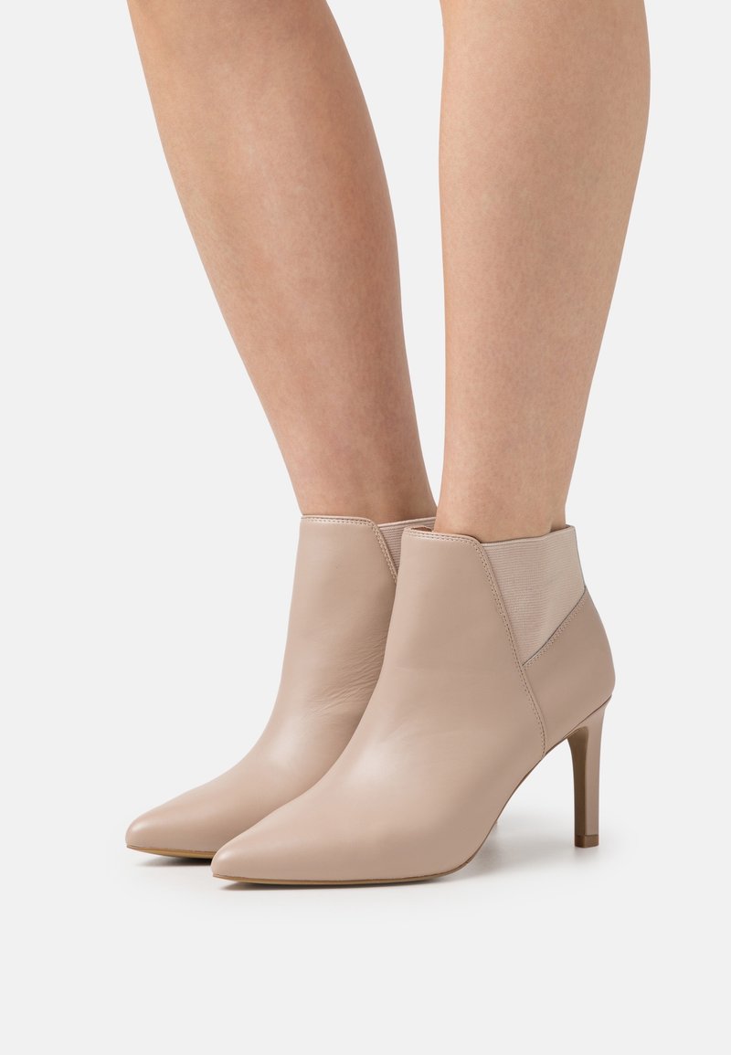 Stivali alla caviglia in pelle beige con punta affilata, pannelli elastici laterali e tacco sottile. Texture liscia con elementi di design minimalisti.