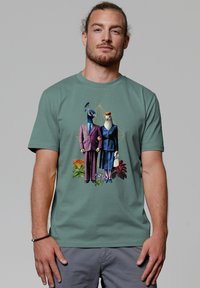 Grafische t-shirt met een illustratie van twee vogels in formele kleding, tegen een groene achtergrond met bloemaccenten.
