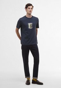 T-shirt de algodão azul-marinho com bolso de retalho às riscas multicoloridas. Combinado com jeans escuros de ganga e mocassins pretos.