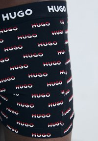 Schwarze Boxer Shorts mit wiederholten roten und weißen "HUGO"-Logo-Mustern. Weiches, elastisches Material mit einem bequemen Bund.