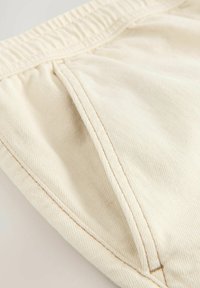 Tissu en coton beige avec une texture lisse, comportant une poche latérale, une taille élastique et des détails de couture contrastés.