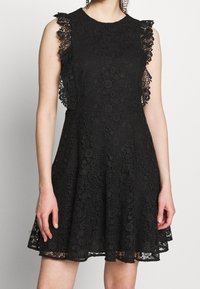 Musta pitsimekko, jossa on tiukka vartalo ja levenevä hame, jossa on röyhelöisiä pitsikoristeita hartioilla ja monimutkaisia kukkakuvioita.