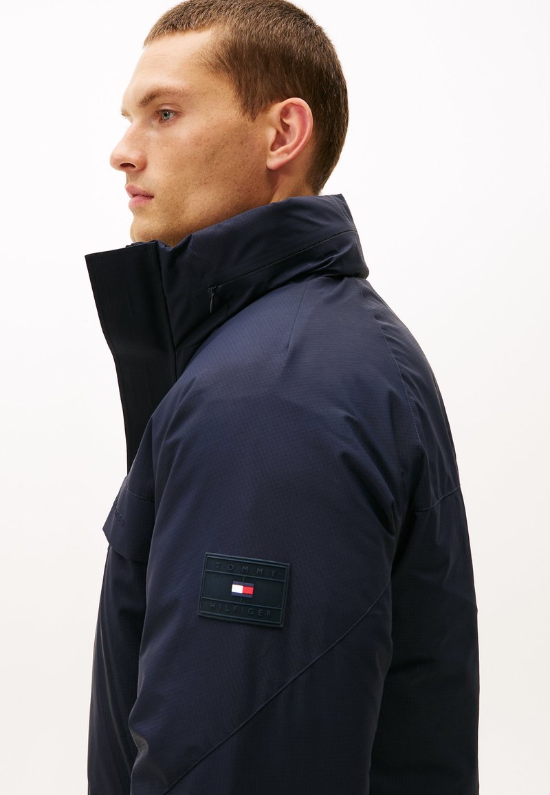 Marinblå vadderad jacka med hög krage, som har en strukturerad yta, dragkedjestängning och en märkespatch på vänster ärm.