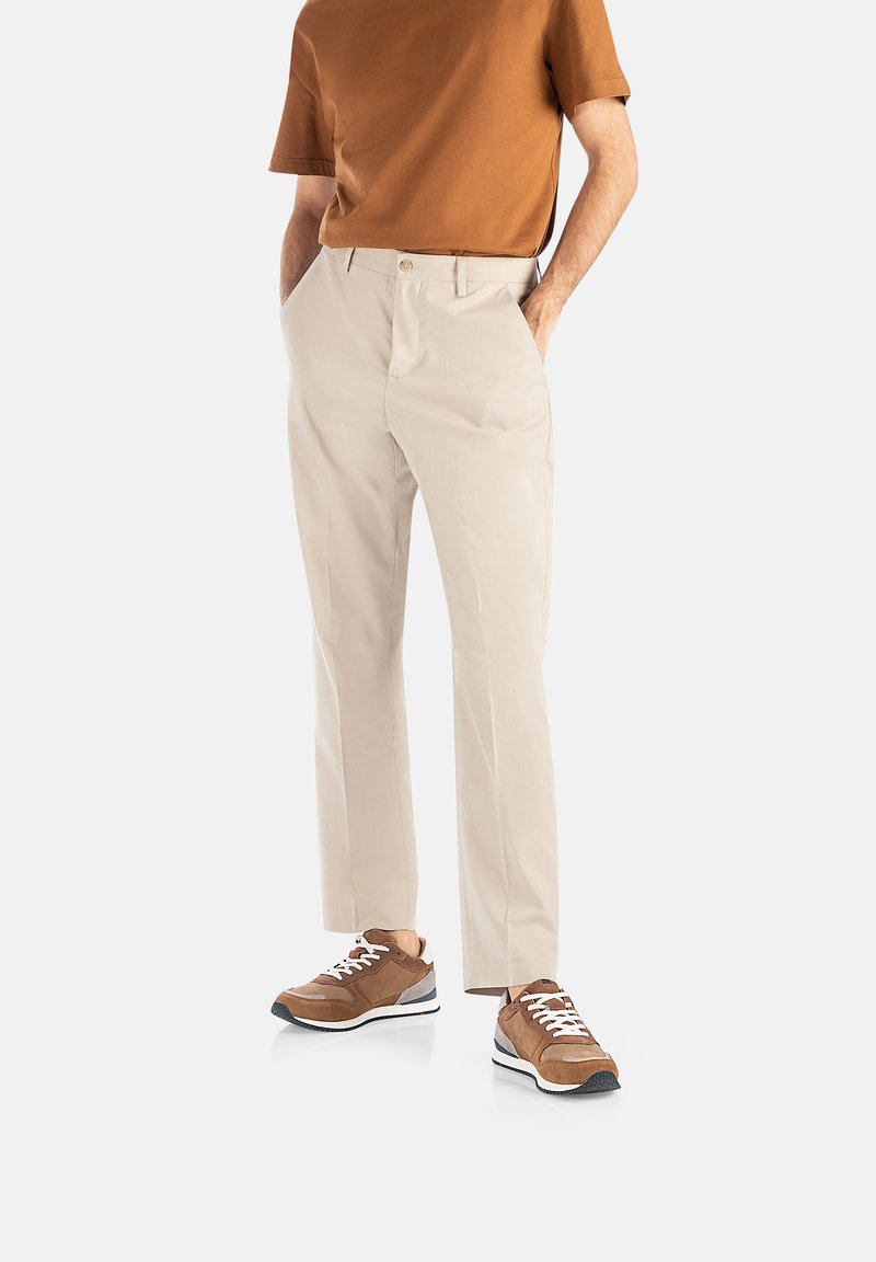 Chinos beige con un corte ajustado, que presentan bolsillos laterales, un cierre frontal con botón y zapatillas deportivas marrones con acentos en contraste.