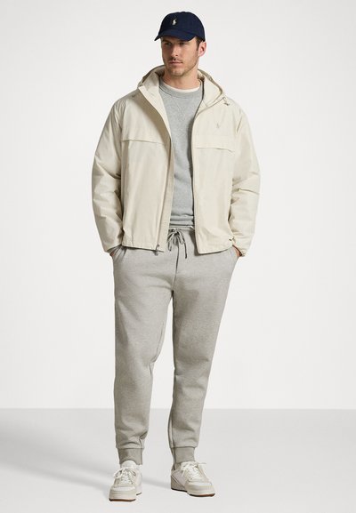 Jachetă bej deschis cu glugă, hanorac gri, pantaloni de trening gri deschis și adidași albi. Prezentare cu logo pe capac și buzunare frontale cu fermoar.