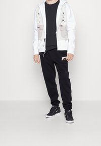 Muž oblečený v černých teniskách, černých teplácích s grafikou, černém tričku a bílé mikině na zip s velkými předními kapsami, stojící na šedé podlaze.